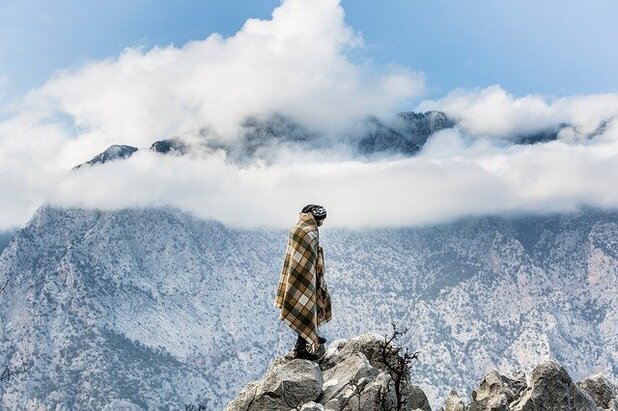 Photo of a nomad, wrapped in a brown-and-tan checked blanket and alone, picking their way across boulders against a backdrop of a mountaintop dusted in snow, capped by wispy white clouds and a blue sky. Courtesy of Pixabay.com.